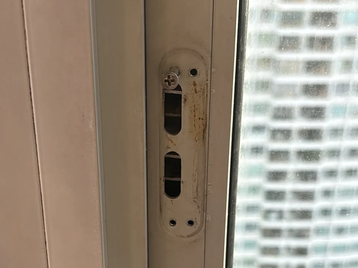 Oval keeper plate recessed into an aluminium window frame, with two slot openings and rust staining